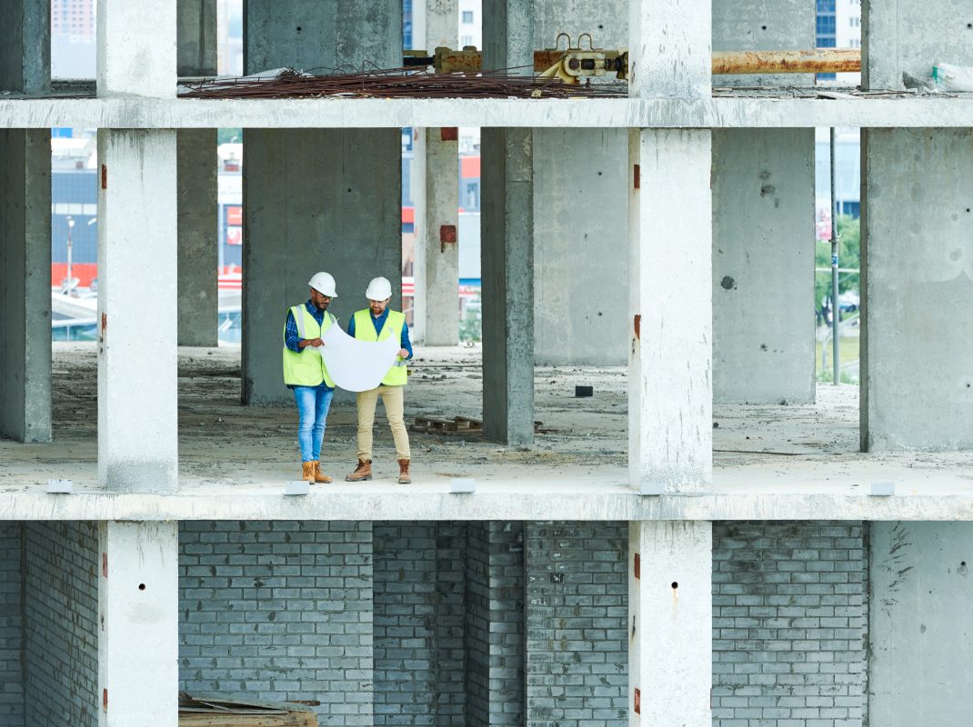 Wide angle view at two builders holding plans standing in unfinished apartment building on construction site, copy space